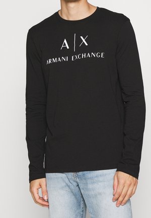 Czarna koszula z długim rękawem z logo "A|X Armani Exchange" w białym kolorze, noszona z jasnoniebieskimi dżinsami, na tle jednolitego jasnego tła.