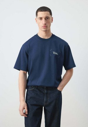 Joven de pie con una mano en el bolsillo, vistiendo una camiseta azul marino con el texto "Yes." y vaqueros oscuros sobre un fondo liso.