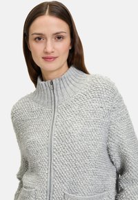 Hellgrauer Zip-Up-Pullover mit hohem gerippten Kragen, der ein strukturiertes Strickmuster und zwei Fronttaschen aufweist. Weiches, gemütliches Material.