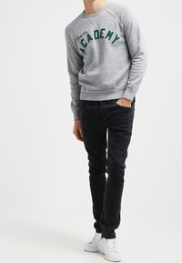 Felpa grigia con la scritta "ACADEMY" in lettere verdi, abbinata a jeans neri. Il modello indossa sneakers bianche, mostrando un look casual.