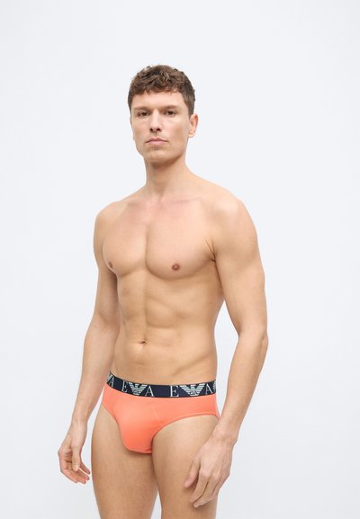 Γυμνασμένος άνδρας με κοντά σγουρά μαλλιά, φοράει πορτοκαλί εσώρουχα με μαύρη μέση που έχει λευκά λογότυπα EA, στέκεται μπροστά σε λευκό, μονόχρωμο φόντο.