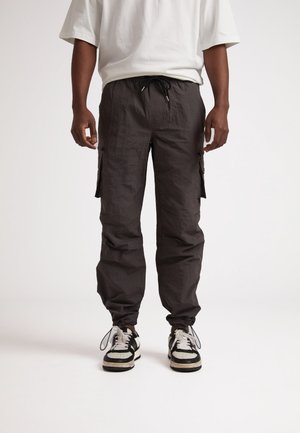 Pantalones cargo oscuros con un acabado texturizado, que cuentan con bolsillos laterales, cintura con cordón y puños elásticos, combinados con zapatillas en blanco y negro.