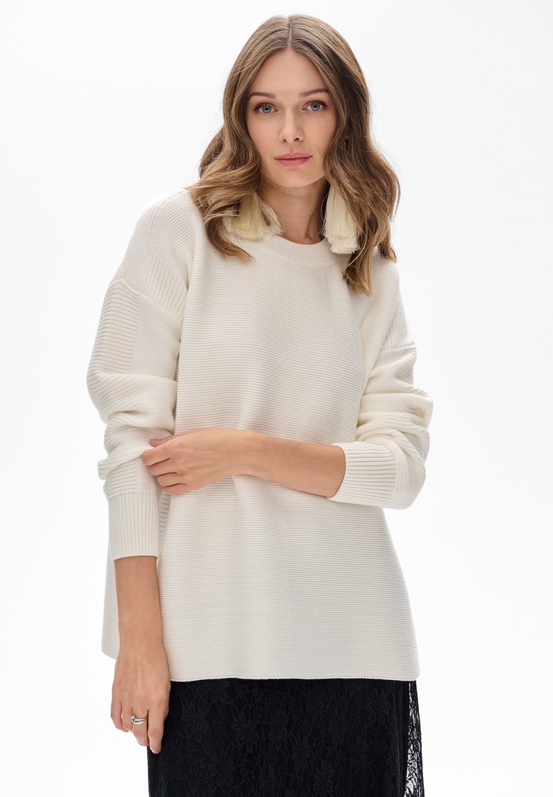 Kobieta z falującymi brązowymi włosami nosi kremowy, prążkowany sweter, duże kremowe kolczyki z frędzlami i czarną koronkową spódnicę, stojąc na tle białym.