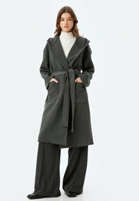 Lang, grijs, wollen blend mantel met brede revers en een strik taille. Bevat twee voorzakken en lange mouwen, gedragen over een witte coltrui en brede broek.