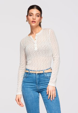 Vrouw met een doorschijnende witte top met patroon en lange mouwen, een beige riem en blauw hoge taille spijkerbroek, staand tegen een effen lichte achtergrond.