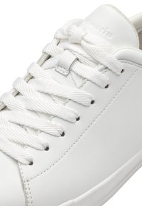 Hvit lær sneakers med flate hvite snøringer, jevn tekstur, minimalistisk design og små svarte hull nær snøringene.
