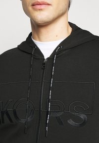 Felpa nera con zip e coulisse, caratterizzata da un logo ricamato di grande rilievo con una texture liscia. Design semplice con vestibilità comoda.