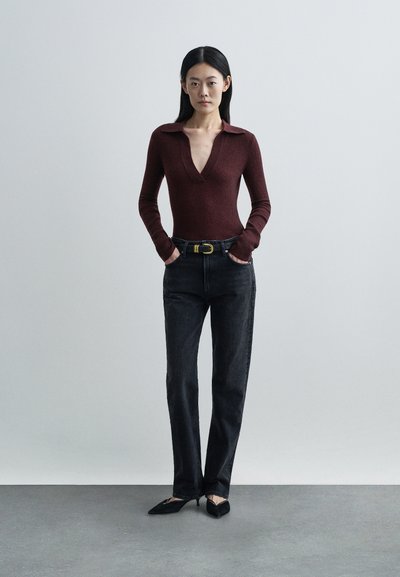 Γυναίκα που στέκεται με τα χέρια στις τσέπες, φορώντας μπορντό μακρυμάνικη μπλούζα, μαύρο τζιν με ζώνη και μαύρες μυτερές γόβες σε γκρι πάτωμα.