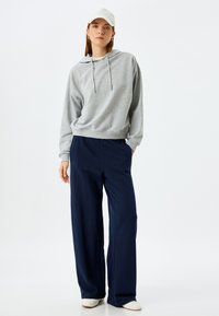 Grijze hoodie met een trekkoord, gecombineerd met marineblauwe wijde broek. De outfit heeft een relaxte pasvorm en wordt gedragen met een witte pet en lichte schoenen.