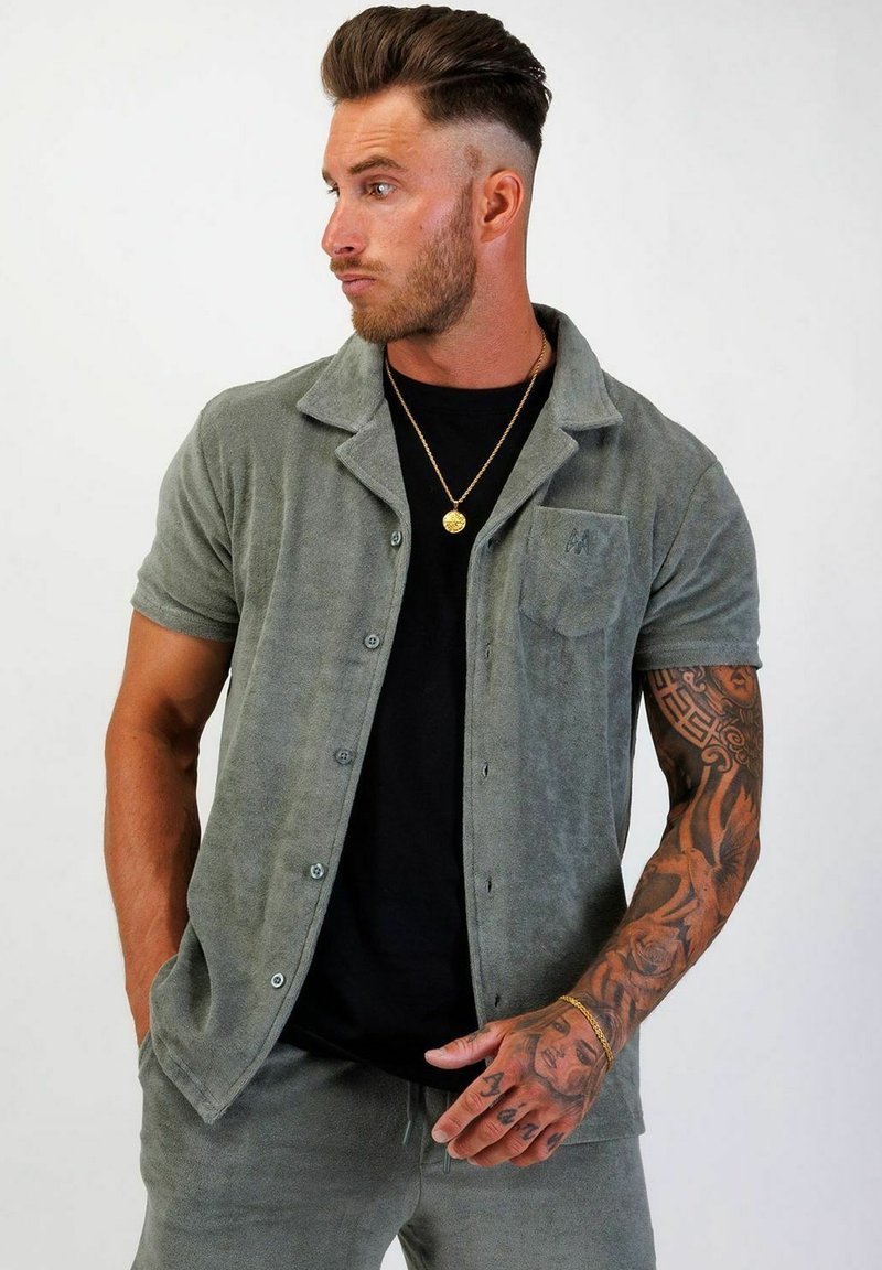 Grön texturerad kortärmad skjorta med knapp-up design, featuring a bröstficka och bärs över en svart T-shirt. Guldig kedja synlig.
