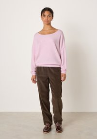 Moteris stovi, dėvi laisvą šviesiai rožinį megztinį, rudas kordurojo kelnes ir rudas odines batus su rožinėmis raištelėmis ant vientisos baltos fono.