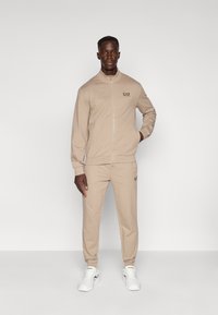 Bēšas krāsas zipa tipa sporta tērps no mīksta auduma, ar piegulošu dizainu un EA7 logotipu uz krūtīm un kājas. Kombinēts ar baltām sporta kurpēm.