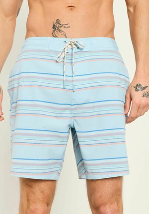 Mann trägt hellblaue gestreifte Badeshorts mit weißem Kordelzug, zeigt Oberkörper sowie Tattoos an Armen und unterem Bauch.