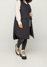 Gilet matelassé noir avec motif en chevrons, présentant des boutons et des poches latérales, porté par-dessus un pull léger et associé à un jean foncé et des bottes volumineuses.