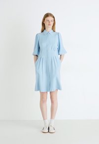 Robe bleu clair à manches courtes, avec un devant boutonné, des plis doux et des poches latérales. Une paire de chaussettes blanches et de baskets complète le look.