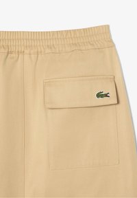 Kaki shorts med en elastisk midja och en bakficka. Har en Lacoste krokodil-logotyp broderad i grönt och rött. Slät textur.