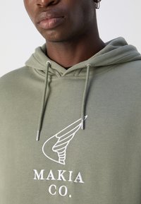 Hoodie vert olive en tissu doux, arborant un motif de logo brodé blanc avec une motif d'ailes et des cordons au niveau de la capuche.