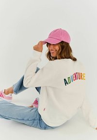 Bílá mikina s multibarevným nápisem "ADVENTURE" na zadní straně, spárovaná se světlými džínami a růžovou baseballovou čepicí a botami.