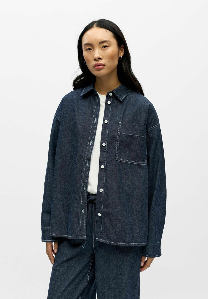 Donkerblauw denim overhemd in een oversized model met een zak, knoopsluiting en witte stikselaccenten. Gecombineerd met een wit T-shirt eronder.