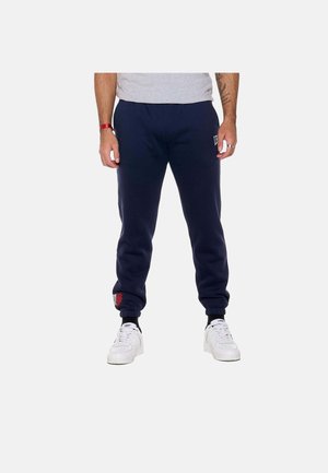 Man draagt marineblauwe joggerbroek met elastische manchetten, witte sneakers en een lichtgrijs shirt, staand tegen een effen achtergrond.