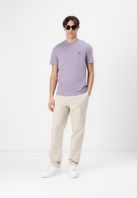 T-shirt lavande avec un petit logo jaune, associé à un pantalon beige et des baskets blanches. Le mannequin porte des lunettes de soleil noires. Design simple, coupe décontractée.