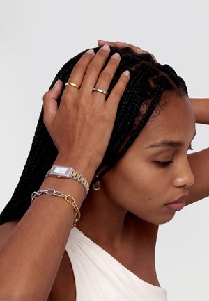 Ženska z pletenimi lasmi drži glavo z obema rokama, nosi zlate in srebrne prstane, dvobarvno uro ter večplastne verižne zapestnice.