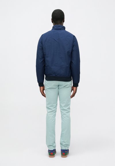 Μπουφάν μπουφάν ναυτικού μπλε με ψηλό γιακά, με λεία υφή. Φορεμένο πάνω από ανοιχτό πράσινο παντελόνι και πολύχρωμα παπούτσια.