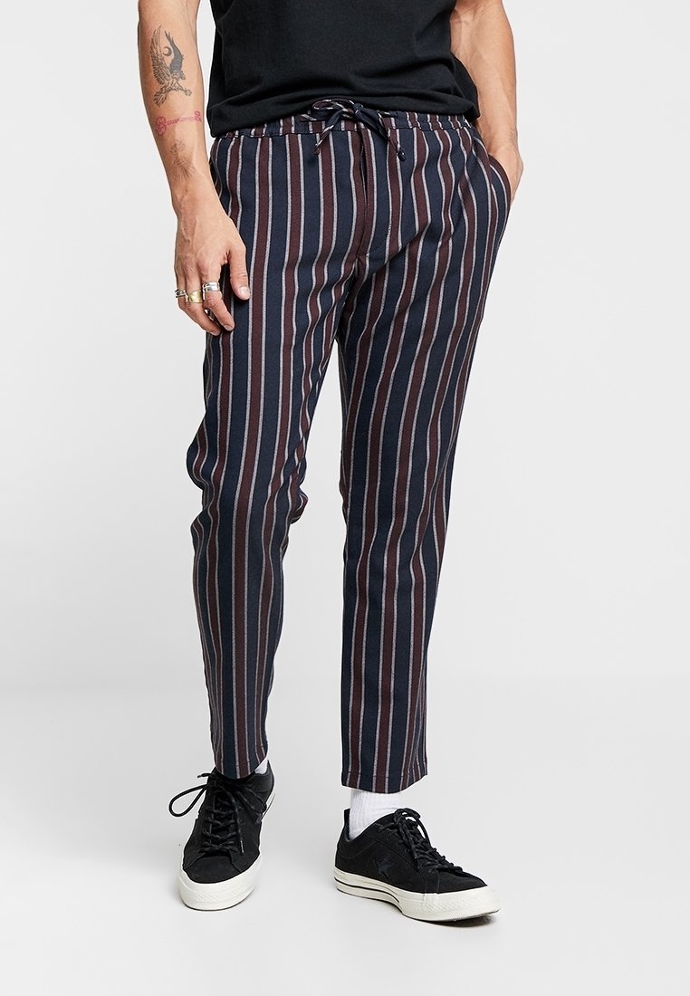 Pantalons rayés avec une taille à cordon en marine foncé, bordeaux et beige. Présentent une coupe slim et des poignets élastiqués, associés à des baskets noires.