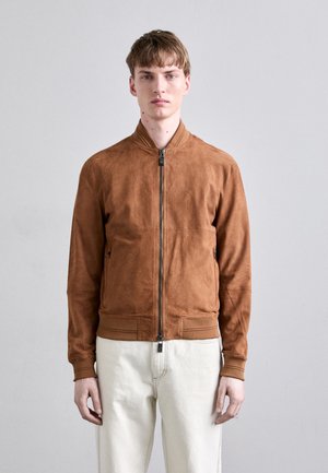 Bomber jacket in suede marrone con chiusura a zip, polsini e orlo a costine, tasche laterali e texture liscia. Indossato sopra pantaloni chiari.