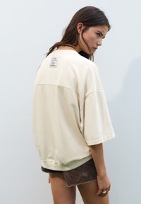 Cremfarbener, übergroßer Pullover mit kurzen Ärmeln, gerippten Bündchen und einem Etikett auf der Rückseite. Kombiniert mit braunen Shorts.