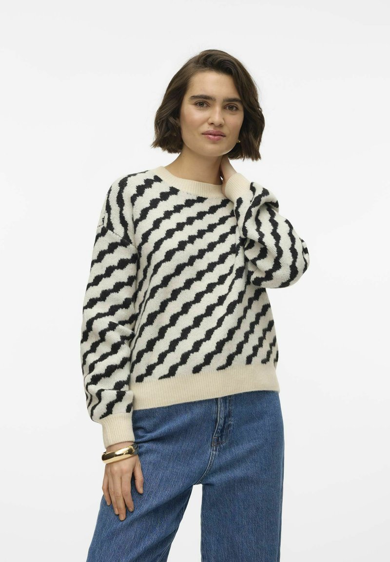 Virkat tröja i krämfärg med svart zigzag-mönster, rund halsringning och sänkta axlar. Kombinerad med hög midja, blå jeans.