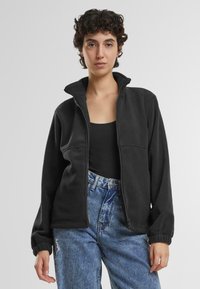 Svart fleece zip-up jakke med høy krage, utstyrt med to frontlommer og tettsittende mansjetter, sammen med høytlivs blå denim jeans.
