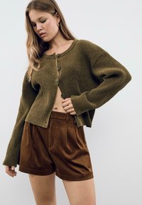 Donna che indossa un cardigan a coste verde oliva con bottoni e pantaloni corti in suede marroni a vita alta, in posa davanti a uno sfondo chiaro e semplice.