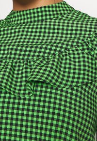 Green and black checkered fabric with a textured ruffle detail along the neckline, featuring a fitted design and high collar.