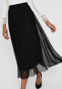 Jupe midi plissée noire avec une taille structurée et un voile transparent. Présente des lignes verticales et une texture fluide, associée à des talons noirs.
