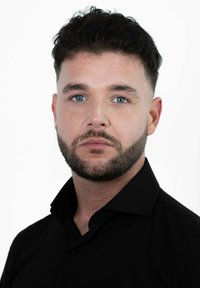 Man met kort krullend donker haar en baard, in een zwart overhemd met kraag, kijkt recht vooruit tegen een witte achtergrond.