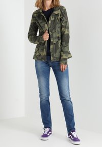 Maskáčová bunda se přiléhavým střihem, předními kapsami a zipem. Oblečená s modrými džíny a teniskami s fialovými akcenty na neutrálním pozadí.