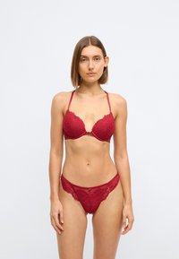 Röd spetsunderkläder set med en djup urringad bh med vågiga kanter och matchande genomskinlig stringtrosa med blommönster och minimal hårdvara.