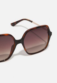 Ochelari de soare supradimensionați, de formă unghiulară, cu rame din fibră de țestoasă și lentile gradient de culoare închisă. Accentele metalice de pe tempă îmbunătățesc designul.