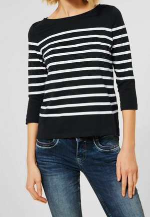 Femme portant un haut à rayures horizontales noires et blanches à manches trois quarts et un jean bleu en denim sur un fond uni.