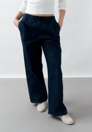 Άτομο που φοράει σκούρο μπλε παντελόνι φαρδιάς γραμμής με ελαστική μέση, λευκά αθλητικά παπούτσια και λευκό μπλουζάκι με μακριά μανίκια, στέκεται με τα χέρια στις τσέπες.