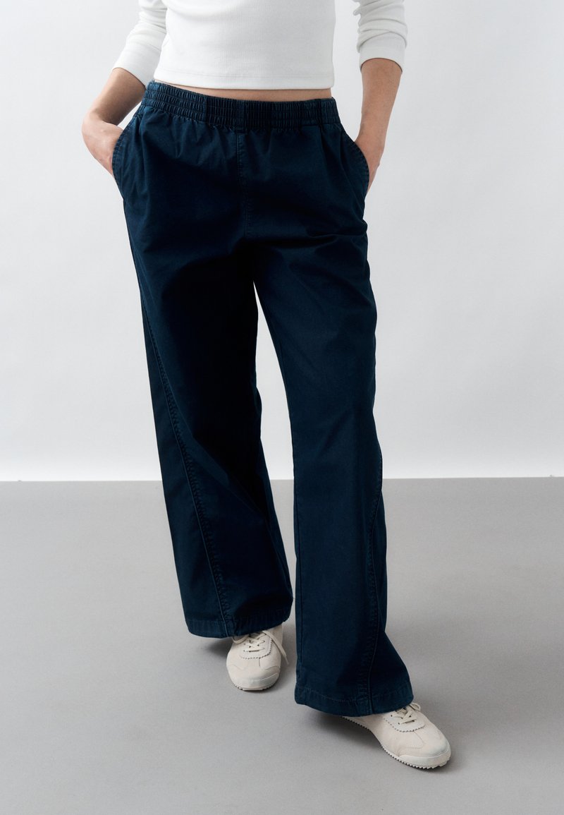 Άτομο που φοράει σκούρο μπλε παντελόνι φαρδιάς γραμμής με ελαστική μέση, λευκά αθλητικά παπούτσια και λευκό μπλουζάκι με μακριά μανίκια, στέκεται με τα χέρια στις τσέπες.