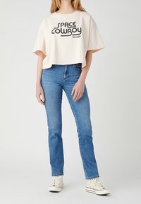 Avklippt, ljusrosa bomullströja med svart "SPACE COWBOY" grafik och logotyp. Bärs med medelblåjeans och vita sneakers.