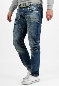 Jeans en denim bleu au design ajusté, présentant des détails usés, des motifs de couture uniques et plusieurs poches. Portés avec des baskets blanches.