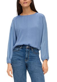 Hellblaues, langärmliges Blusenshirt mit weitem Ausschnitt, fließendem Stoff und leicht gerafften Manschetten, kombiniert mit klassischen blauen Jeans.