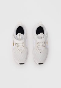 Białe buty do biegania Nike z złotymi logo Swoosh, rzepami i sznurowadłami widziane z góry na jednolitym białym tle.