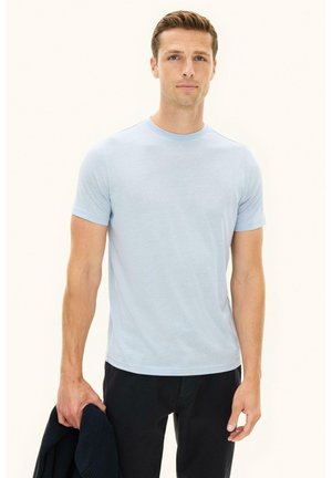 Mann trägt ein hellblaues kurzärmeliges T-Shirt und schwarze Hosen, hält eine dunkle Jacke in der linken Hand und steht vor einem schlichten hellen Hintergrund.