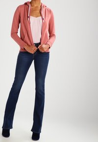 Top sin mangas de color rosa claro debajo de una sudadera con capucha y cremallera de color coral. Jeans acampanados de color azul oscuro. La modelo está de pie, con las manos sosteniendo la sudadera.