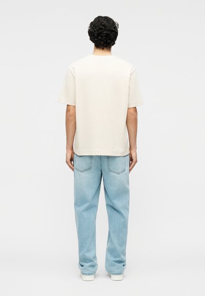 Άνδρας με μαύρα σγουρά μαλλιά στέκεται με την πλάτη, φοράει απλό κρεμ κοντομάνικο πουκάμισο, ανοιχτό μπλε τζιν παντελόνι και λευκά παπούτσια.