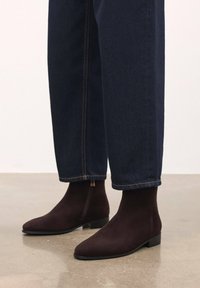 Barna velúr bokacsizmák elegáns, hegyes orral és alacsony blokk sarkal, sötétkék, cropped farmernadrággal kombinálva, narancssárga díszítéssel.